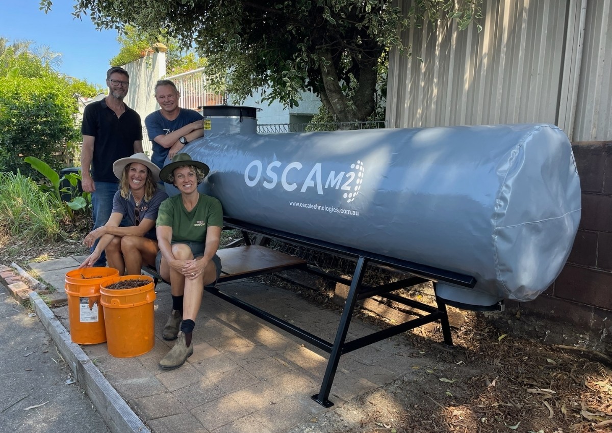 Four people posing with large cylindrical OSCA composter outdoors, two seated with buckets of organic material, demonstrating OSCA's sustainable waste management system and circular economy composting operations