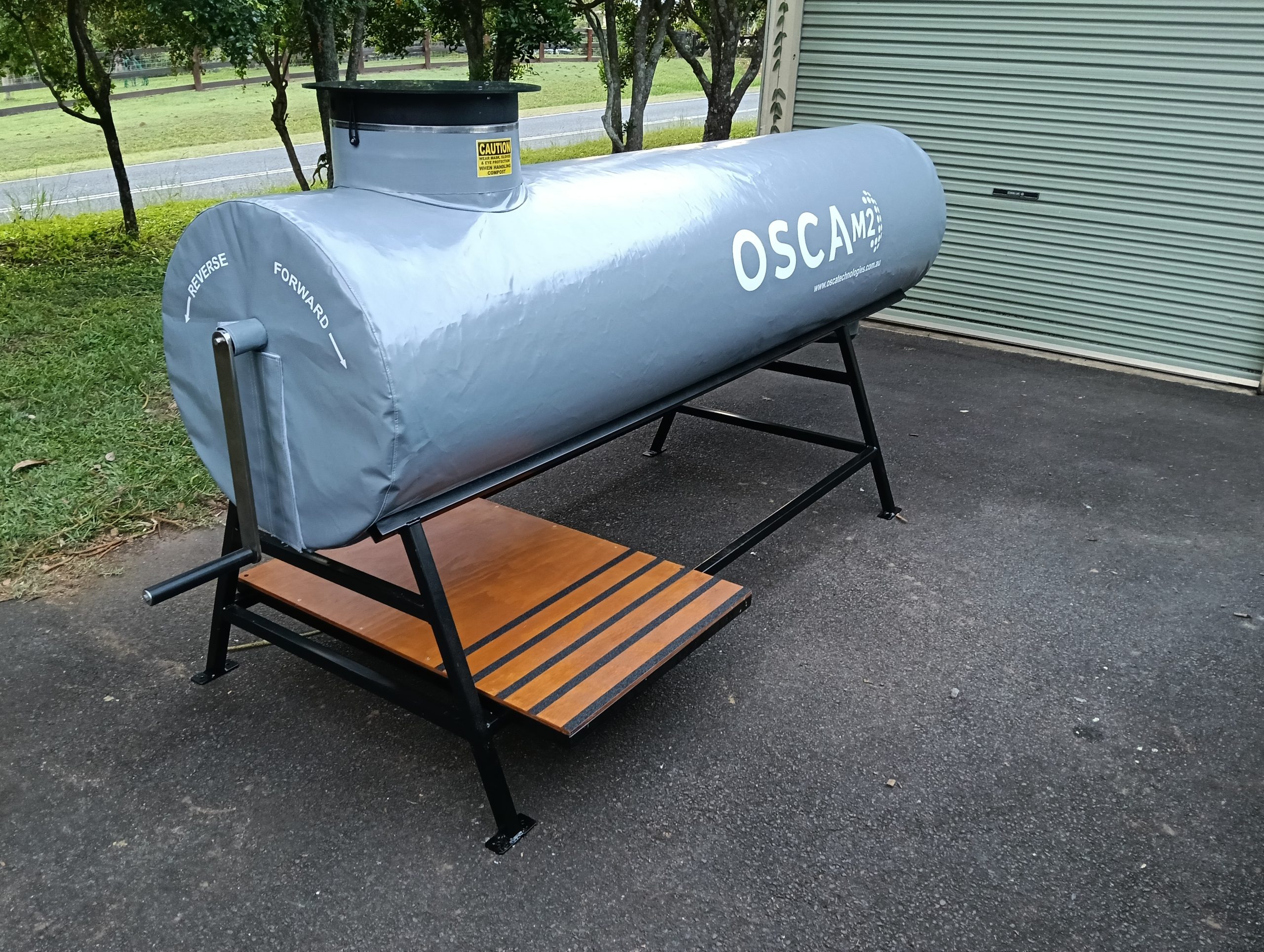 Large grey cylindrical OSCA composter tank with ventilation vent on top, mounted on black metal frame with wooden platform, installed on residential driveway for sustainable organic waste processing and composting operations